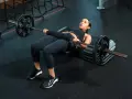Donna esegue un **hamstring bridge** con bilanciere su una panca in palestra, concentrata sull'esercizio.