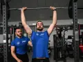 Uomo esegue push press con bilanciere, assistito da un trainer in palestra.