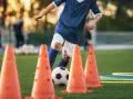 Giovane calciatore si allena con esercizi rapidità calcio senza palla, slalom tra i coni arancioni su un campo verde.