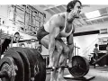 Arnold Schwarzenegger esegue uno stiff legged deadlift in palestra, concentrato sul sollevamento del bilanciere.