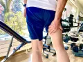 Uomo con mano sulla coscia, indicando una possibile lesione hamstring. Attrezzatura da palestra e alberi fioriti sullo sfondo.