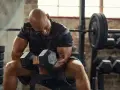 Uomo calvo in palestra, concentrato su un curl con manubri da 50 kg. Il suo viso mostra lo sforzo, indicando un RPE chart elevato.