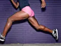 Gambe muscolose di un corridore che esegue esercizi per i posteriori della coscia, con sfondo di mattoni viola.
