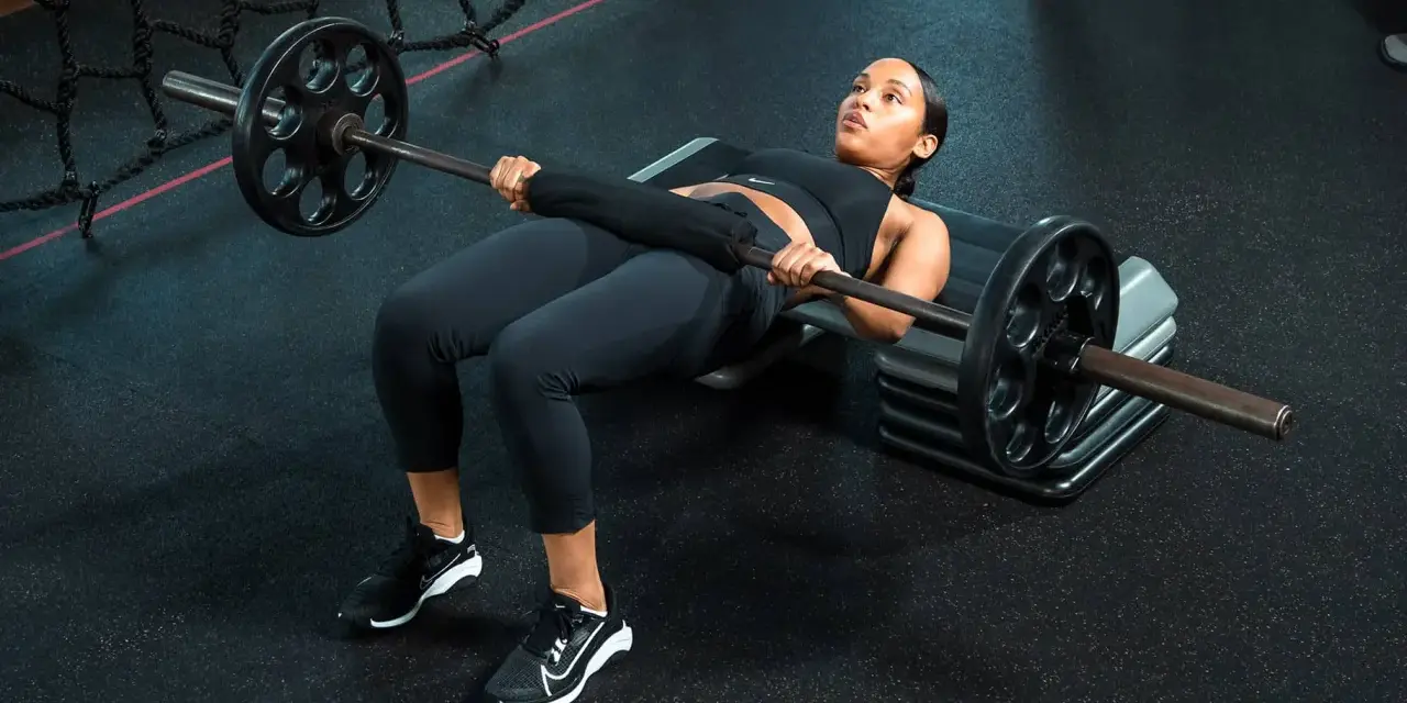 Donna esegue un **hamstring bridge** con bilanciere su una panca in palestra, concentrata sull'esercizio.