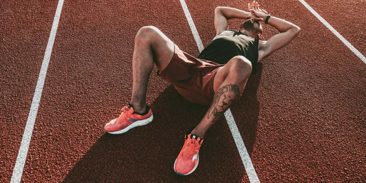 Atleta esausto a terra su una pista da corsa, mani sulla testa. Potrebbe essere il **significato di overtraining**: il corpo che chiede una pausa.