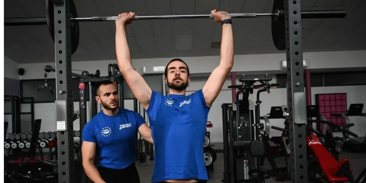 Uomo esegue push press con bilanciere, assistito da un trainer in palestra.