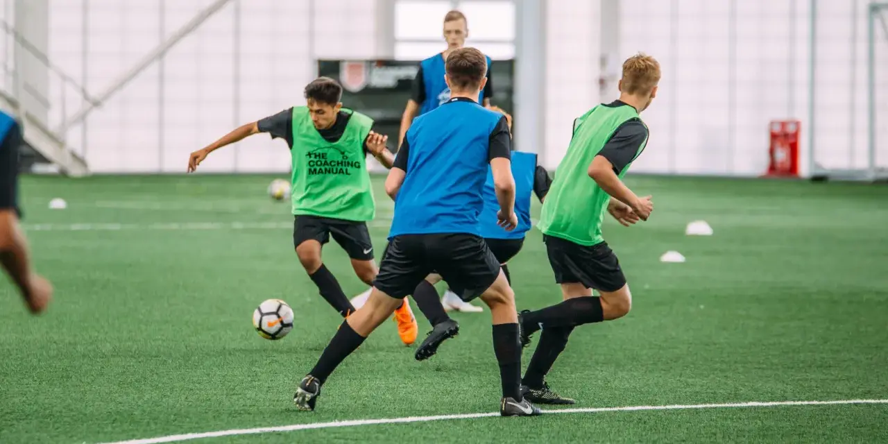 Giovani calciatori in allenamento, con pettorine verdi e blu, corrono su un campo sintetico. L'intensità dell'allenamento richiede un **aerobico alto**.