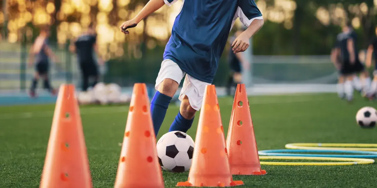 Giovane calciatore si allena con esercizi rapidità calcio senza palla, slalom tra i coni arancioni su un campo verde.