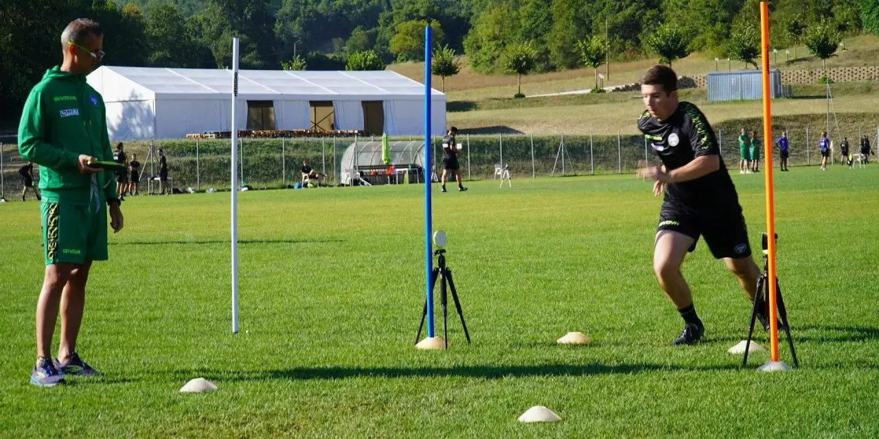 Allenatore osserva un atleta eseguire un test di agilità, con coni e paletti disposti sul campo per il **yoyo test tabella**.