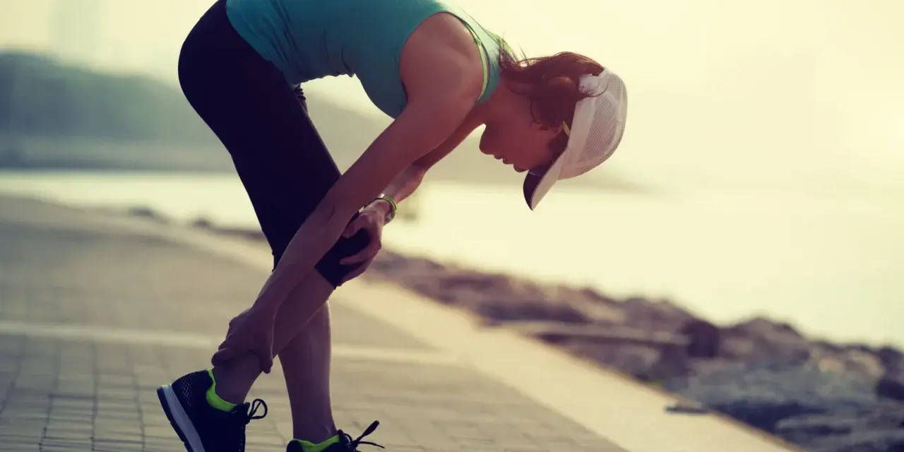 Donna con cappellino si piega tenendosi il ginocchio, forse a causa di un infortunio durante la corsa. La sua espressione suggerisce dolore, ma la sua determinazione è evidente. La scala di Borg potrebbe essere usata per valutare l'intensità dello sforzo.