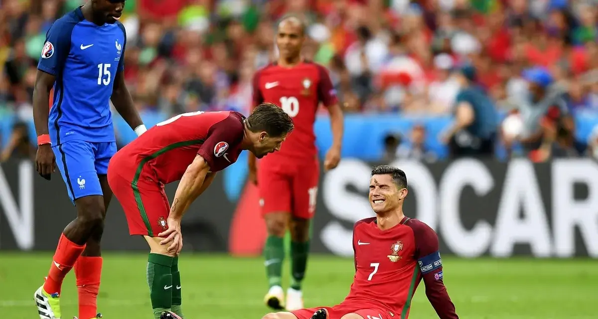 Un giocatore portoghese a terra, forse per un infortunio calcio, mentre un compagno si china preoccupato e un avversario osserva.