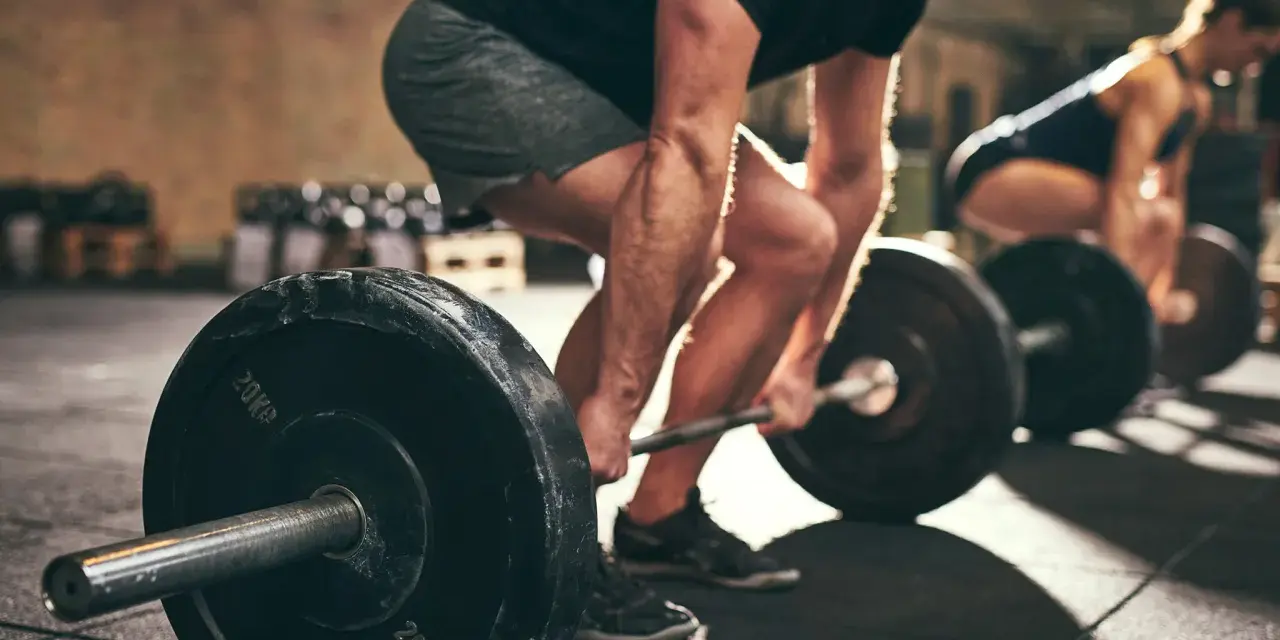 Uomo e donna si allenano in palestra con bilancieri. I suoi **doms bicipiti** sono visibili mentre solleva il peso.