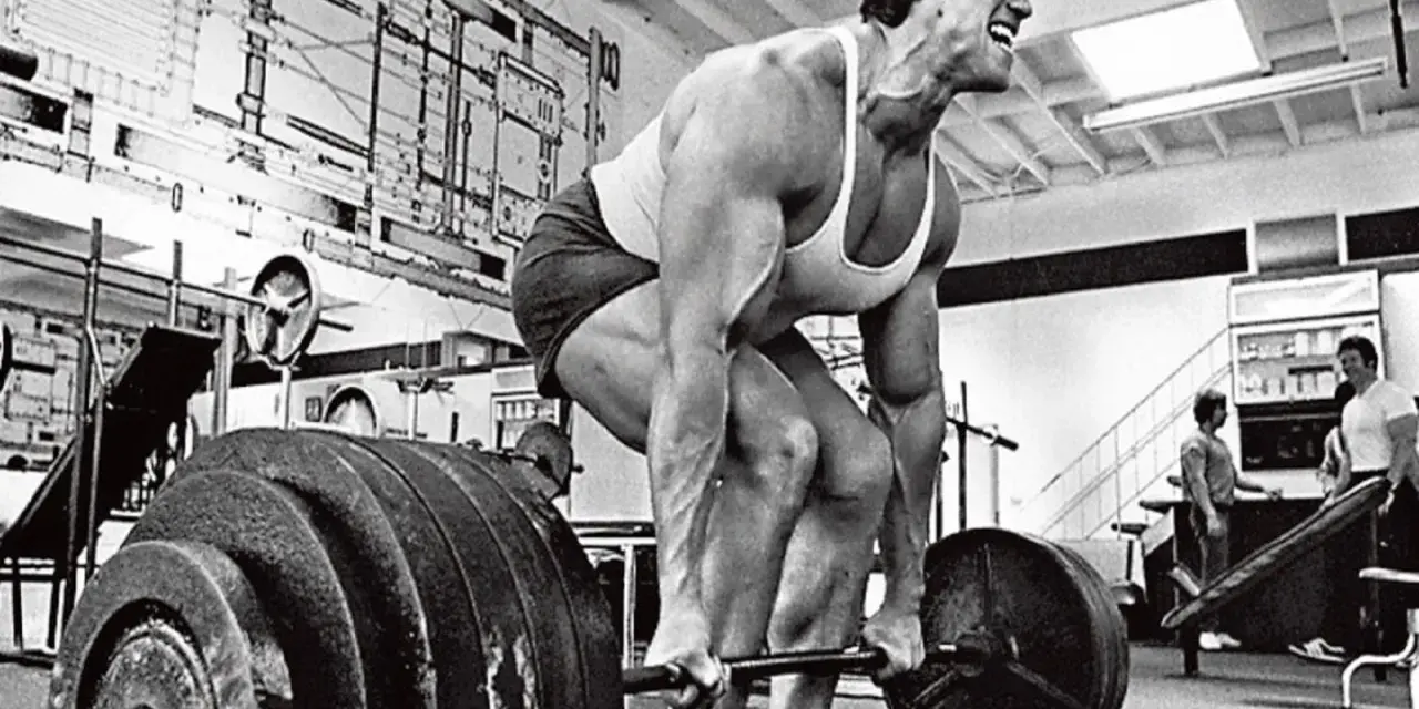 Arnold Schwarzenegger esegue uno stiff legged deadlift in palestra, concentrato sul sollevamento del bilanciere.