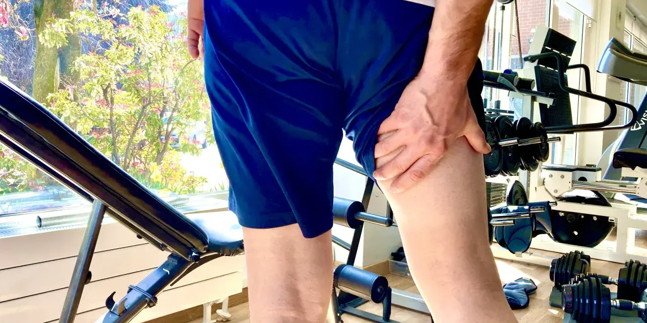 Uomo con mano sulla coscia, indicando una possibile lesione hamstring. Attrezzatura da palestra e alberi fioriti sullo sfondo.