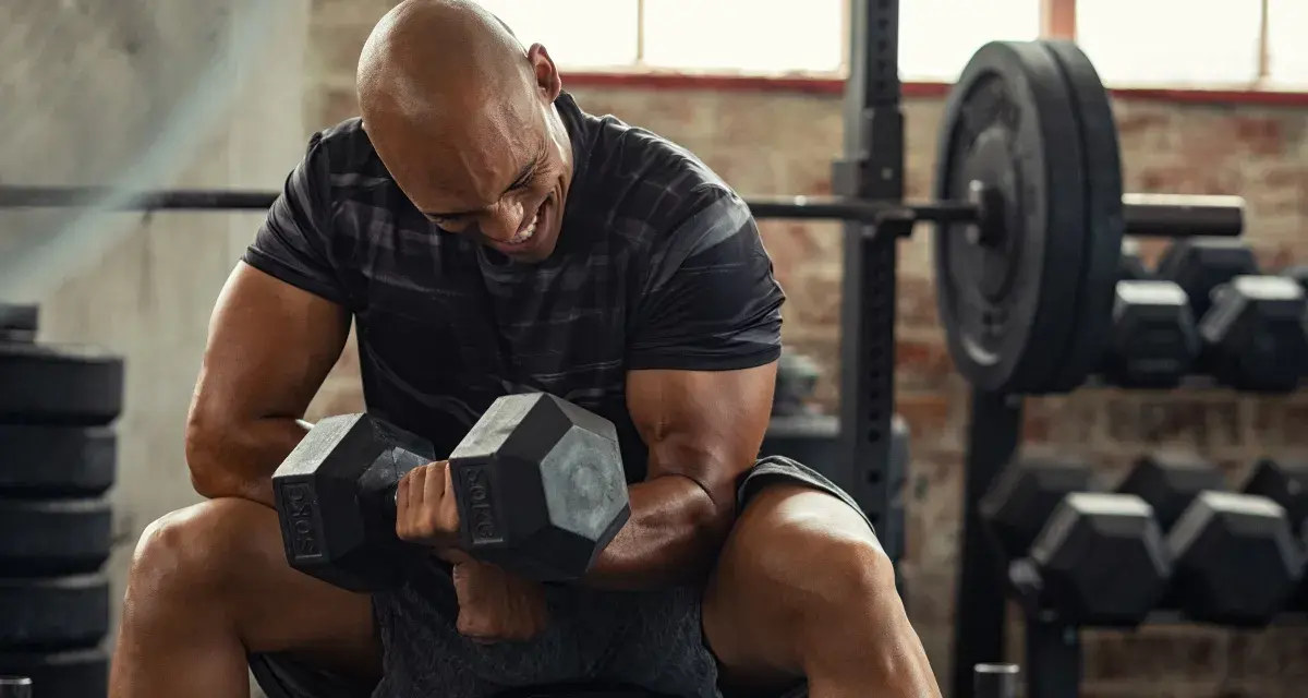 Uomo calvo in palestra, concentrato su un curl con manubri da 50 kg. Il suo viso mostra lo sforzo, indicando un RPE chart elevato.