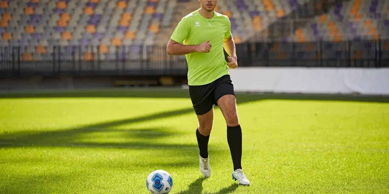 Giovane calciatore corre sul campo verde, allenando la resistenza. Un pallone da calcio è vicino a lui.