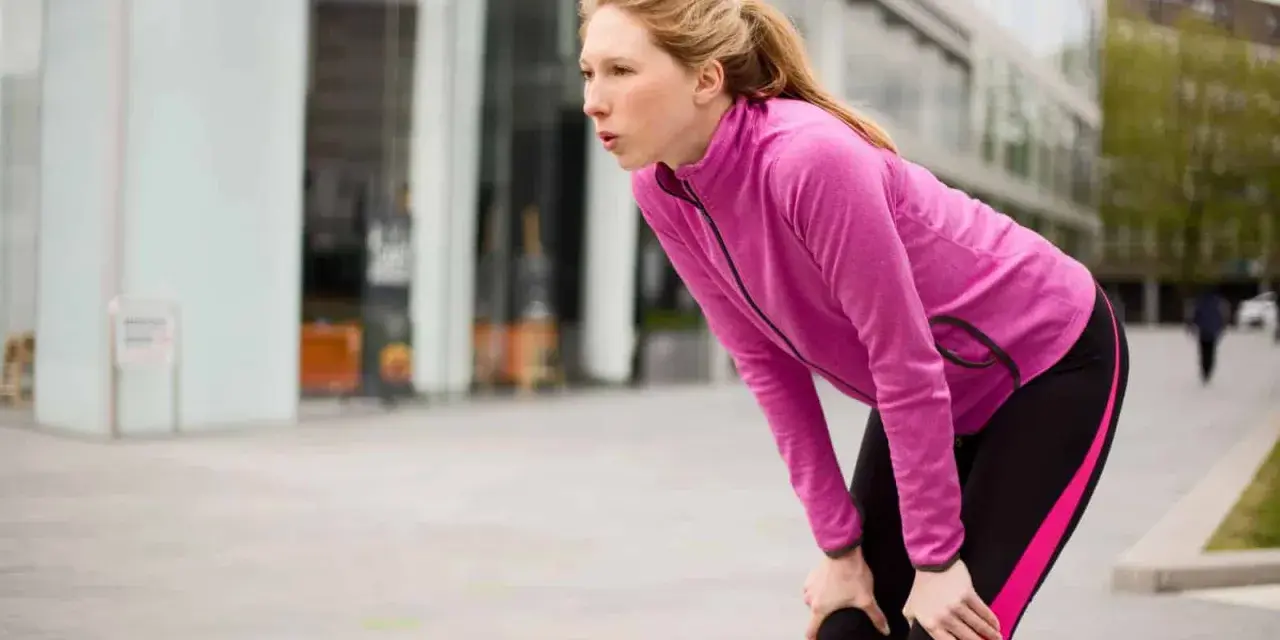 Donna bionda, esausta dopo la corsa, si piega appoggiando le mani sulle ginocchia. Il suo respiro affannoso indica un alto RPE (Rate of Perceived Exertion).
