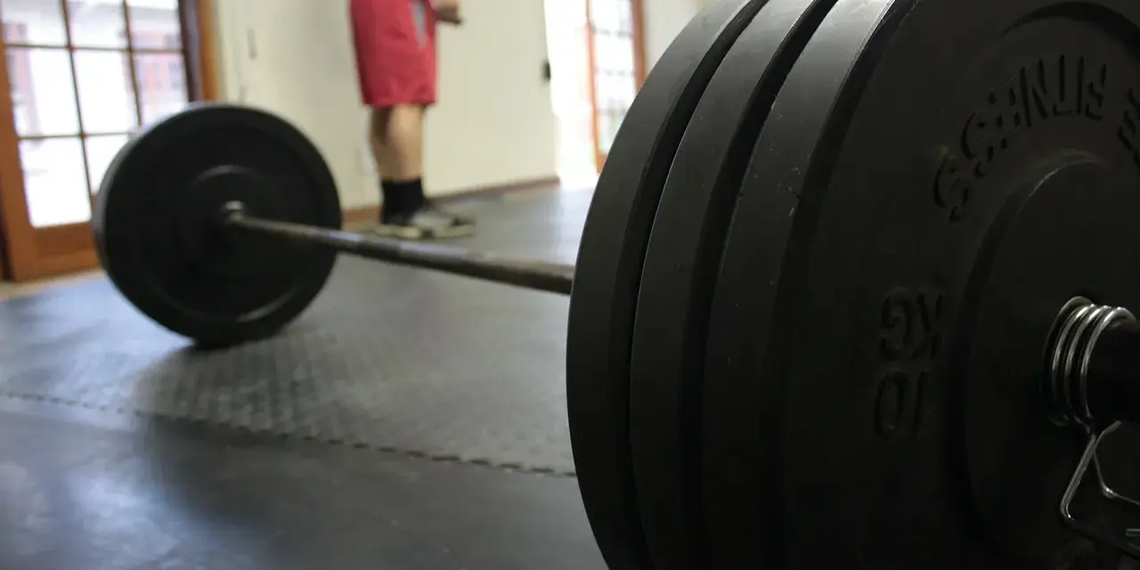 Bilanciere con pesi pronti per un allenamento sulla panca piana. Un uomo in pantaloncini rossi attende sullo sfondo.