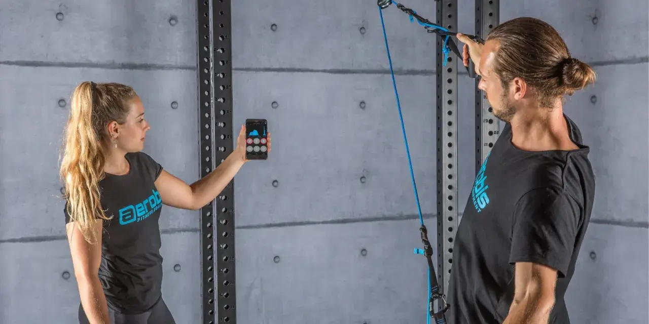 Donna con smartphone e uomo con cavo per allenamento. Il display mostra grafici, forse per misurare la forza o il progresso, spiegando cosa significa pr.