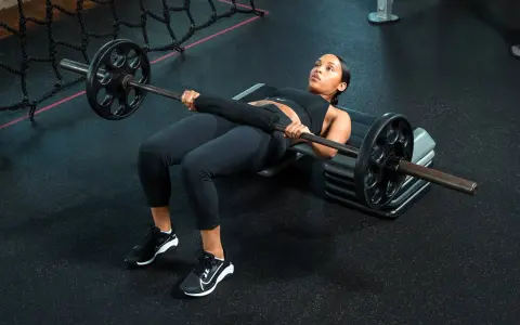 Donna esegue un **hamstring bridge** con bilanciere su una panca in palestra, concentrata sull'esercizio.