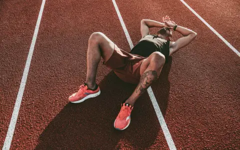 Atleta esausto a terra su una pista da corsa, mani sulla testa. Potrebbe essere il **significato di overtraining**: il corpo che chiede una pausa.