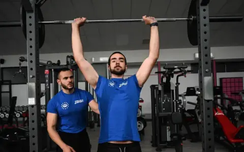 Uomo esegue push press con bilanciere, assistito da un trainer in palestra.