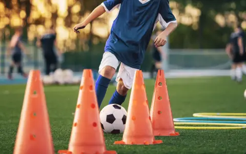 Giovane calciatore si allena con esercizi rapidità calcio senza palla, slalom tra i coni arancioni su un campo verde.