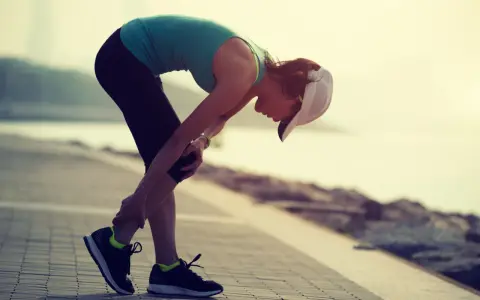 Donna con cappellino si piega tenendosi il ginocchio, forse a causa di un infortunio durante la corsa. La sua espressione suggerisce dolore, ma la sua determinazione è evidente. La scala di Borg potrebbe essere usata per valutare l'intensità dello sforzo.