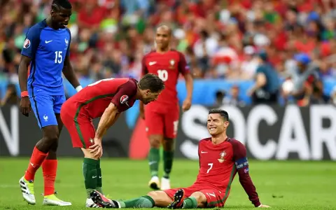 Un giocatore portoghese a terra, forse per un infortunio calcio, mentre un compagno si china preoccupato e un avversario osserva.
