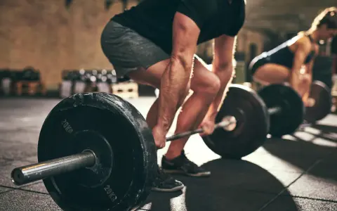 Uomo e donna si allenano in palestra con bilancieri. I suoi **doms bicipiti** sono visibili mentre solleva il peso.