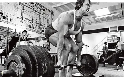 Arnold Schwarzenegger esegue uno stiff legged deadlift in palestra, concentrato sul sollevamento del bilanciere.