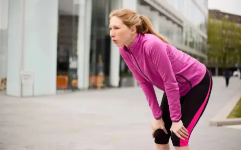 Donna bionda, esausta dopo la corsa, si piega appoggiando le mani sulle ginocchia. Il suo respiro affannoso indica un alto RPE (Rate of Perceived Exertion).