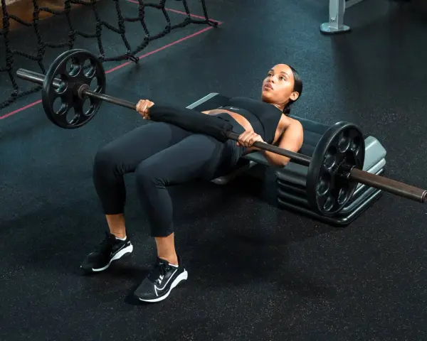 Donna esegue un **hamstring bridge** con bilanciere su una panca in palestra, concentrata sull'esercizio.
