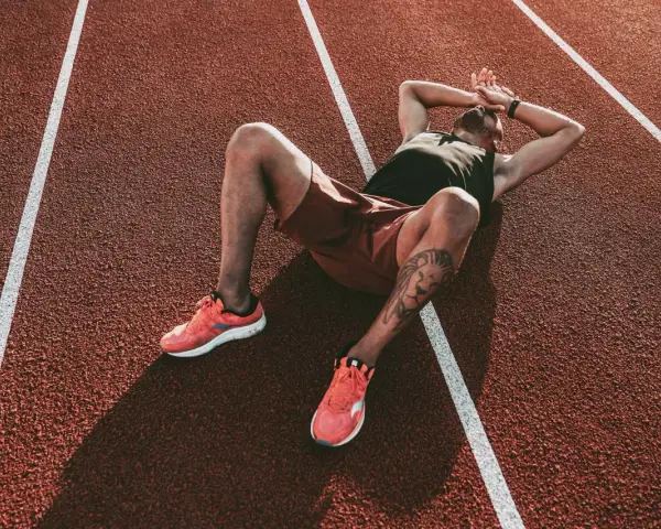 Atleta esausto a terra su una pista da corsa, mani sulla testa. Potrebbe essere il **significato di overtraining**: il corpo che chiede una pausa.