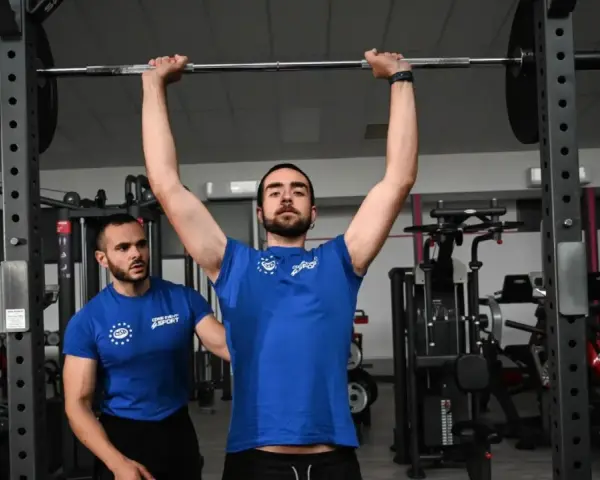 Uomo esegue push press con bilanciere, assistito da un trainer in palestra.