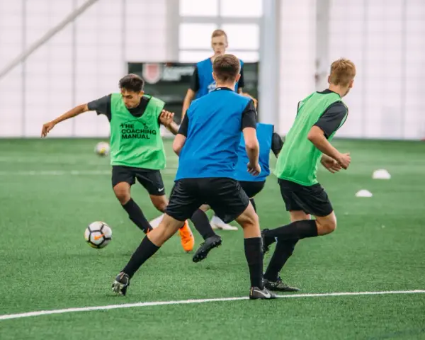 Giovani calciatori in allenamento, con pettorine verdi e blu, corrono su un campo sintetico. L'intensità dell'allenamento richiede un **aerobico alto**.