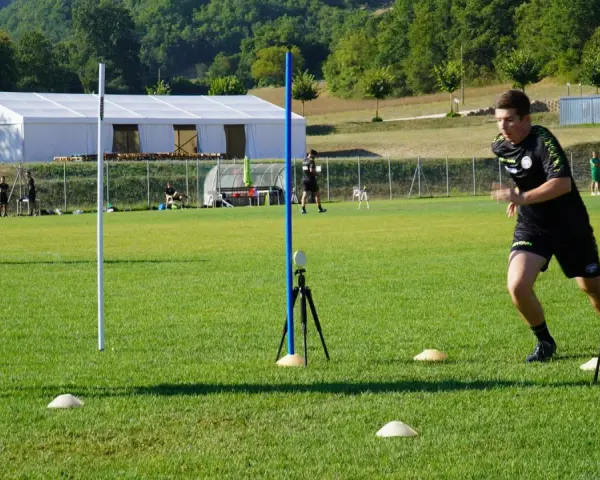 Allenatore osserva un atleta eseguire un test di agilità, con coni e paletti disposti sul campo per il **yoyo test tabella**.