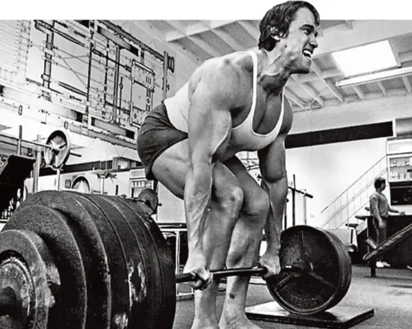 Arnold Schwarzenegger esegue uno stiff legged deadlift in palestra, concentrato sul sollevamento del bilanciere.