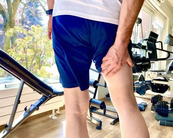 Uomo con mano sulla coscia, indicando una possibile lesione hamstring. Attrezzatura da palestra e alberi fioriti sullo sfondo.