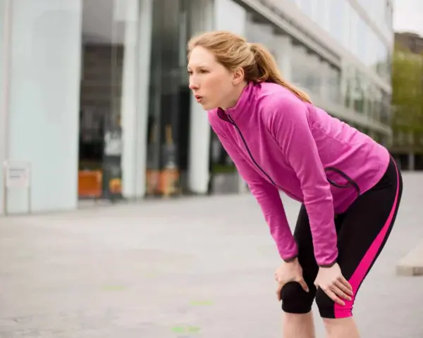Donna bionda, esausta dopo la corsa, si piega appoggiando le mani sulle ginocchia. Il suo respiro affannoso indica un alto RPE (Rate of Perceived Exertion).