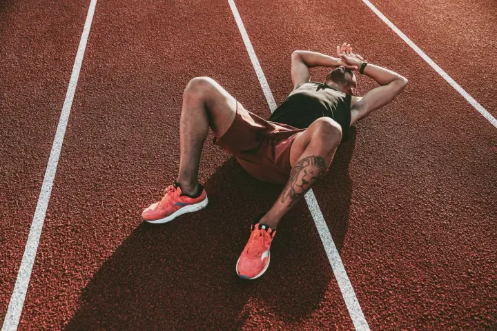 Atleta esausto a terra su una pista da corsa, mani sulla testa. Potrebbe essere il **significato di overtraining**: il corpo che chiede una pausa.