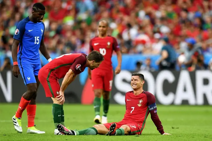 Un giocatore portoghese a terra, forse per un infortunio calcio, mentre un compagno si china preoccupato e un avversario osserva.