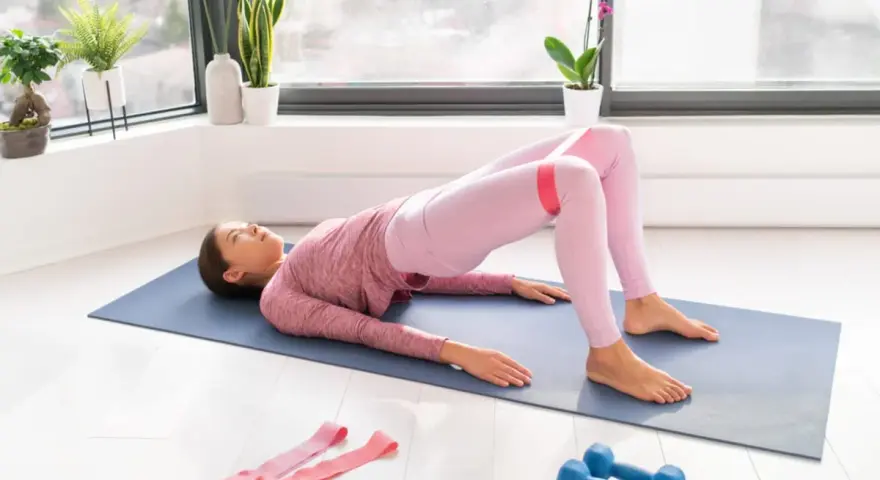 Donna esegue hip thrust a casa con elastico rosa sulle cosce. Sul tappetino, vicino a due manubri blu, si allena per glutei forti.