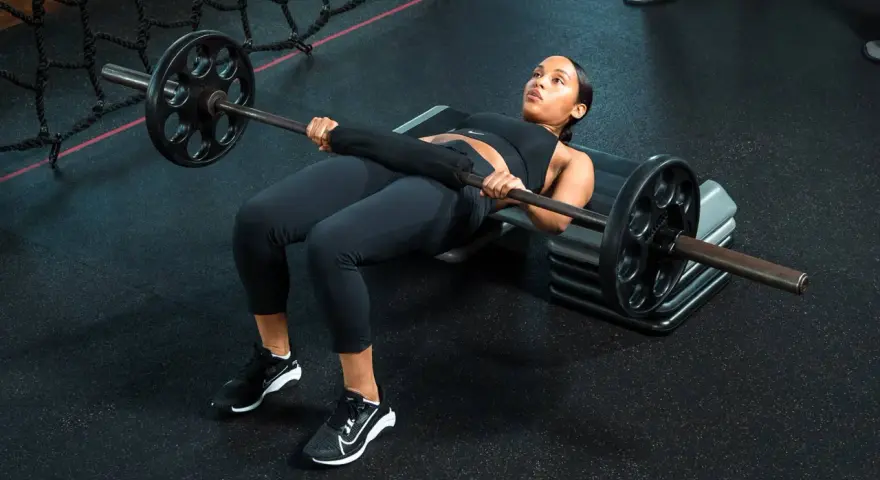 Donna esegue un **hamstring bridge** con bilanciere su una panca in palestra, concentrata sull'esercizio.