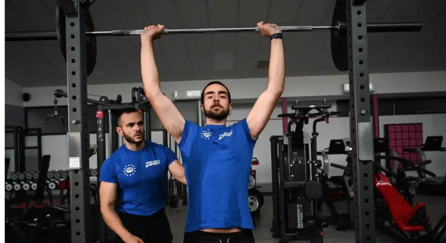 Uomo esegue push press con bilanciere, assistito da un trainer in palestra.