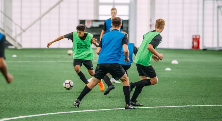 Giovani calciatori in allenamento, con pettorine verdi e blu, corrono su un campo sintetico. L'intensità dell'allenamento richiede un **aerobico alto**.