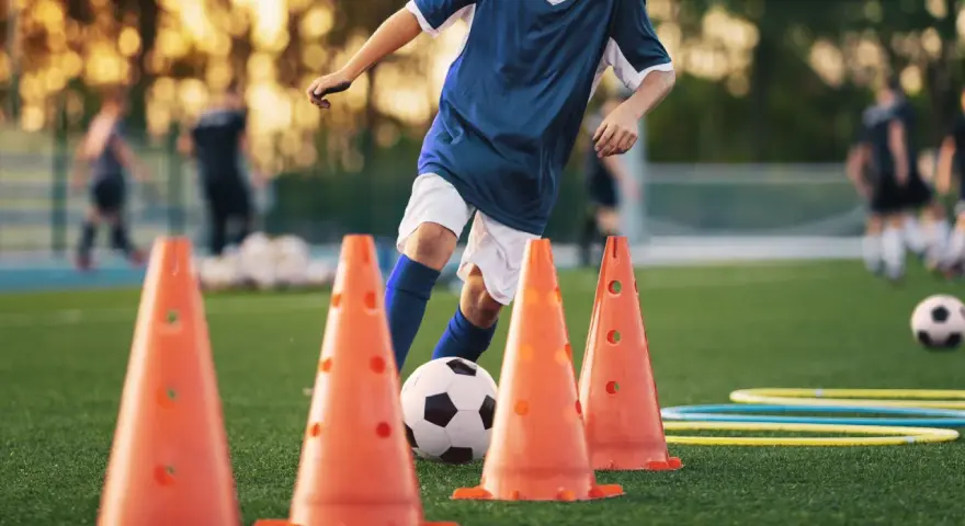 Giovane calciatore si allena con esercizi rapidità calcio senza palla, slalom tra i coni arancioni su un campo verde.