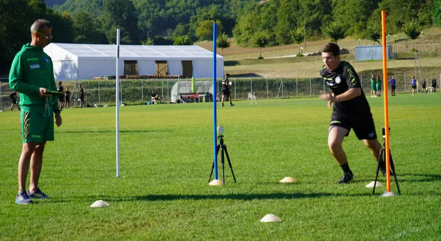 Allenatore osserva un atleta eseguire un test di agilità, con coni e paletti disposti sul campo per il **yoyo test tabella**.