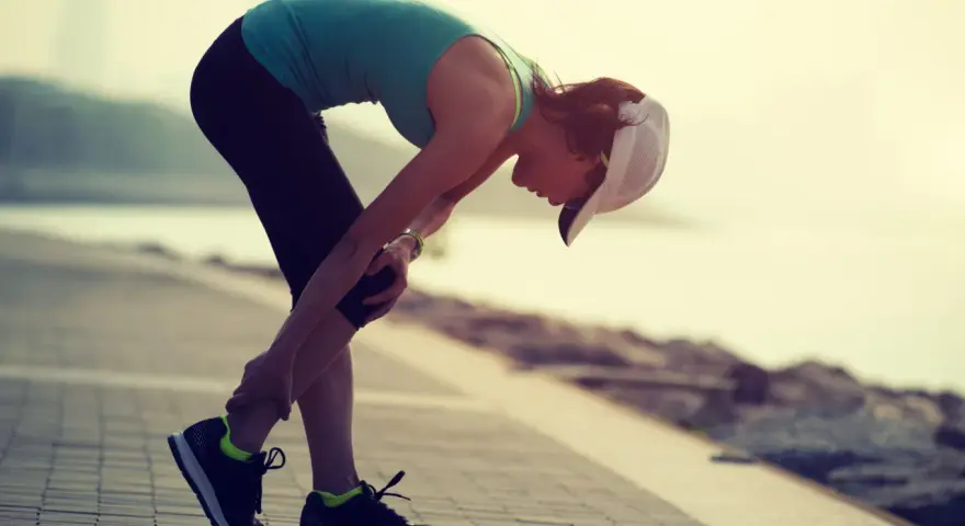 Donna con cappellino si piega tenendosi il ginocchio, forse a causa di un infortunio durante la corsa. La sua espressione suggerisce dolore, ma la sua determinazione è evidente. La scala di Borg potrebbe essere usata per valutare l'intensità dello sforzo.
