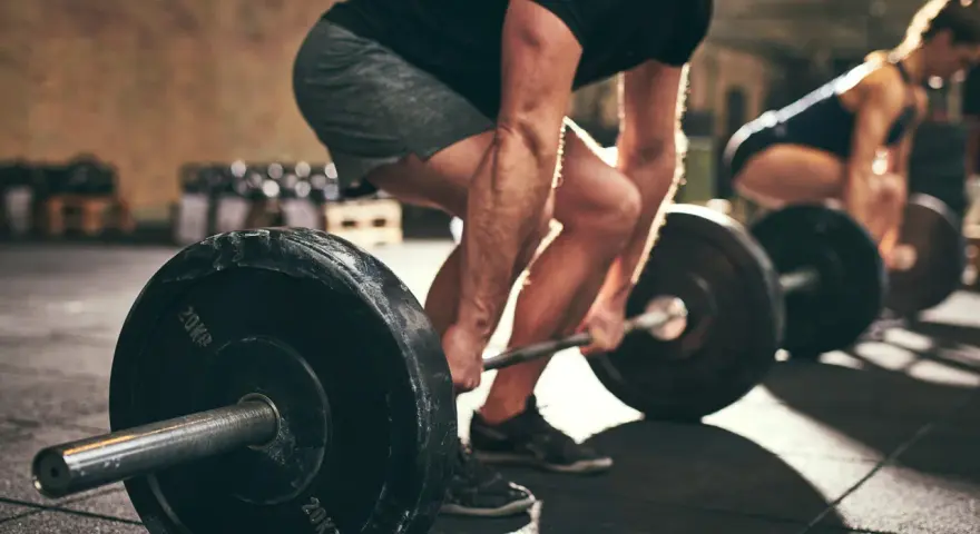 Uomo e donna si allenano in palestra con bilancieri. I suoi **doms bicipiti** sono visibili mentre solleva il peso.